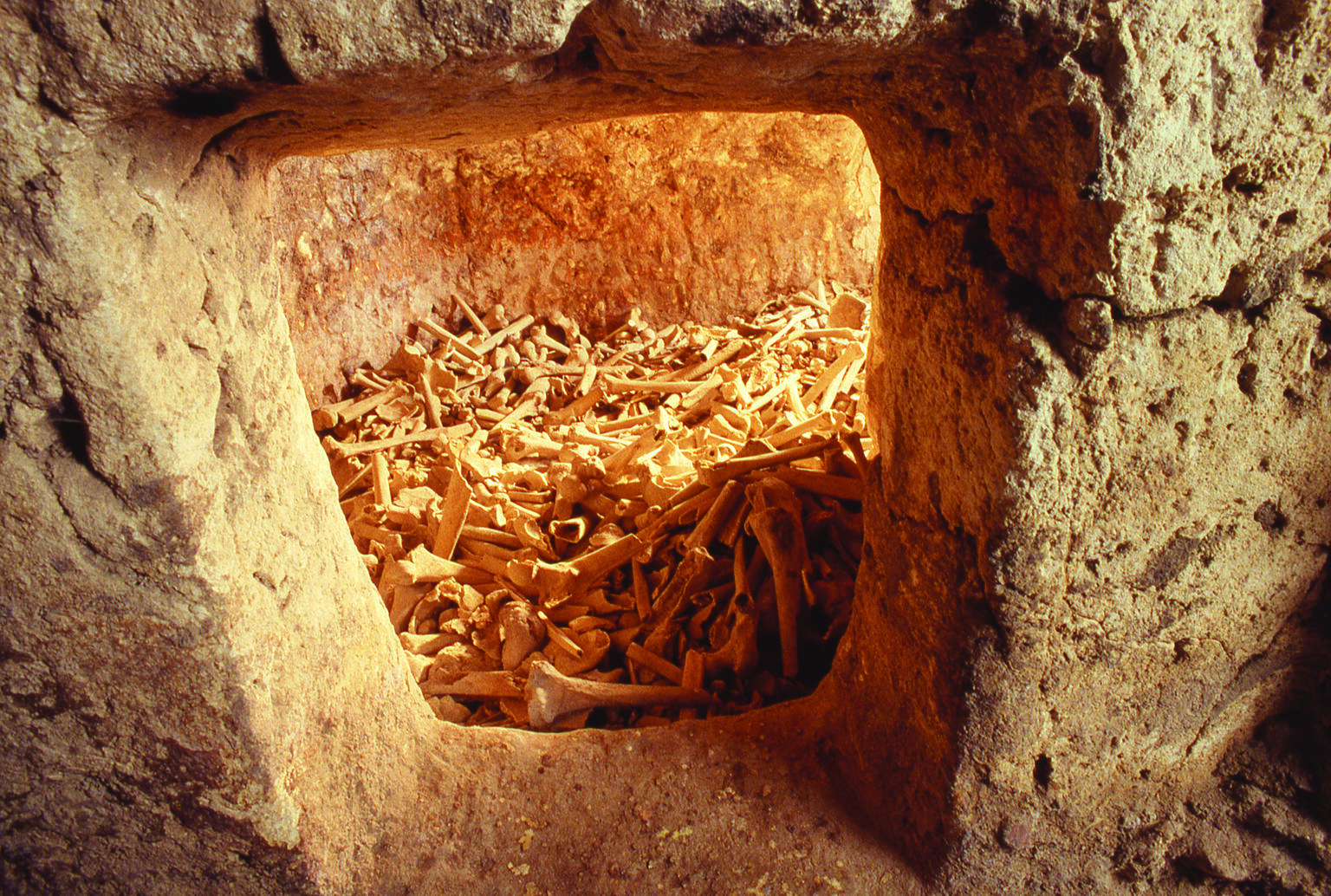 Rectangular window within stone tomb revealing cache of many human bones.