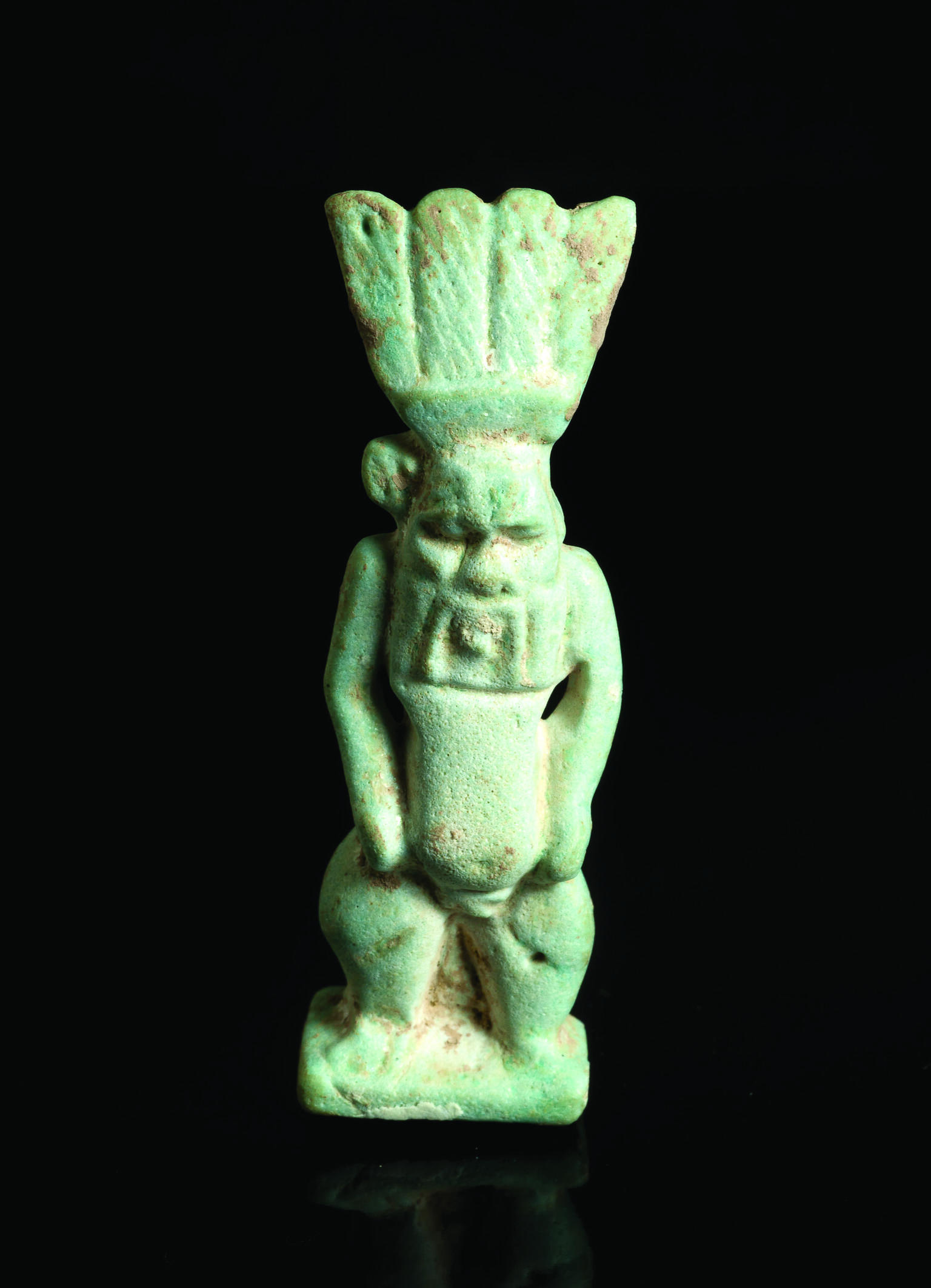 Frontal and back faience amulet of figure who is nude with feathered headdress and a grotesque face and beard.