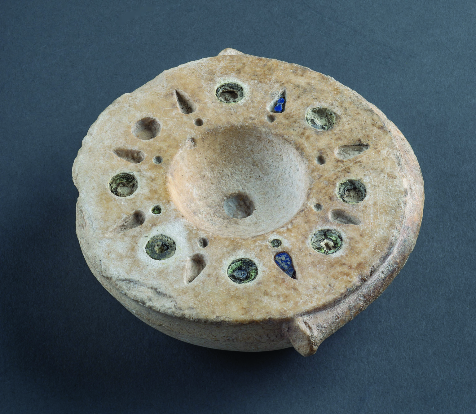 Limestone bowl with mosaic decoration around rim with alternating circular and bud-shaped inlaid blue and brown glass. 