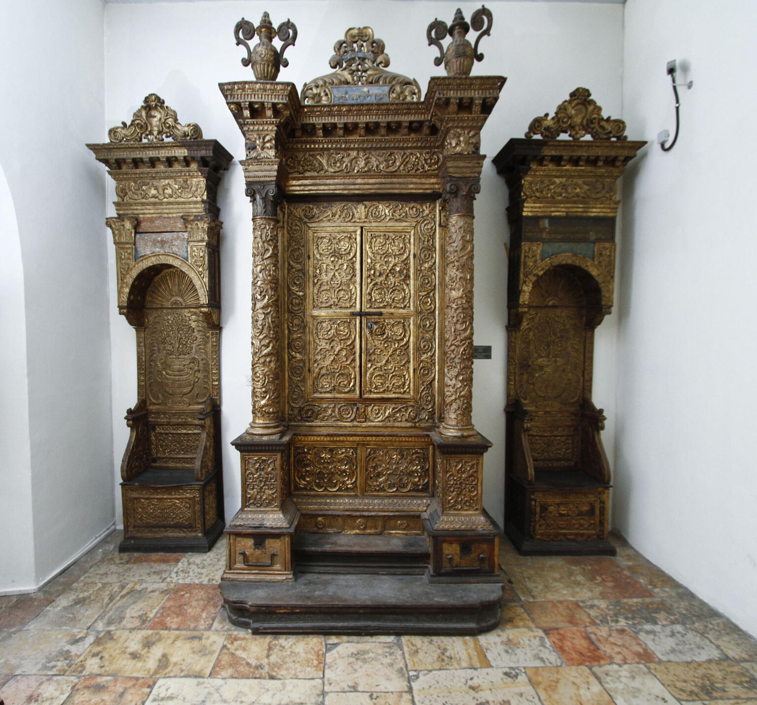 Wooden Torah ark decorated with carvings, columns, and capitals, flanked by two similarly decorated chairs. 
