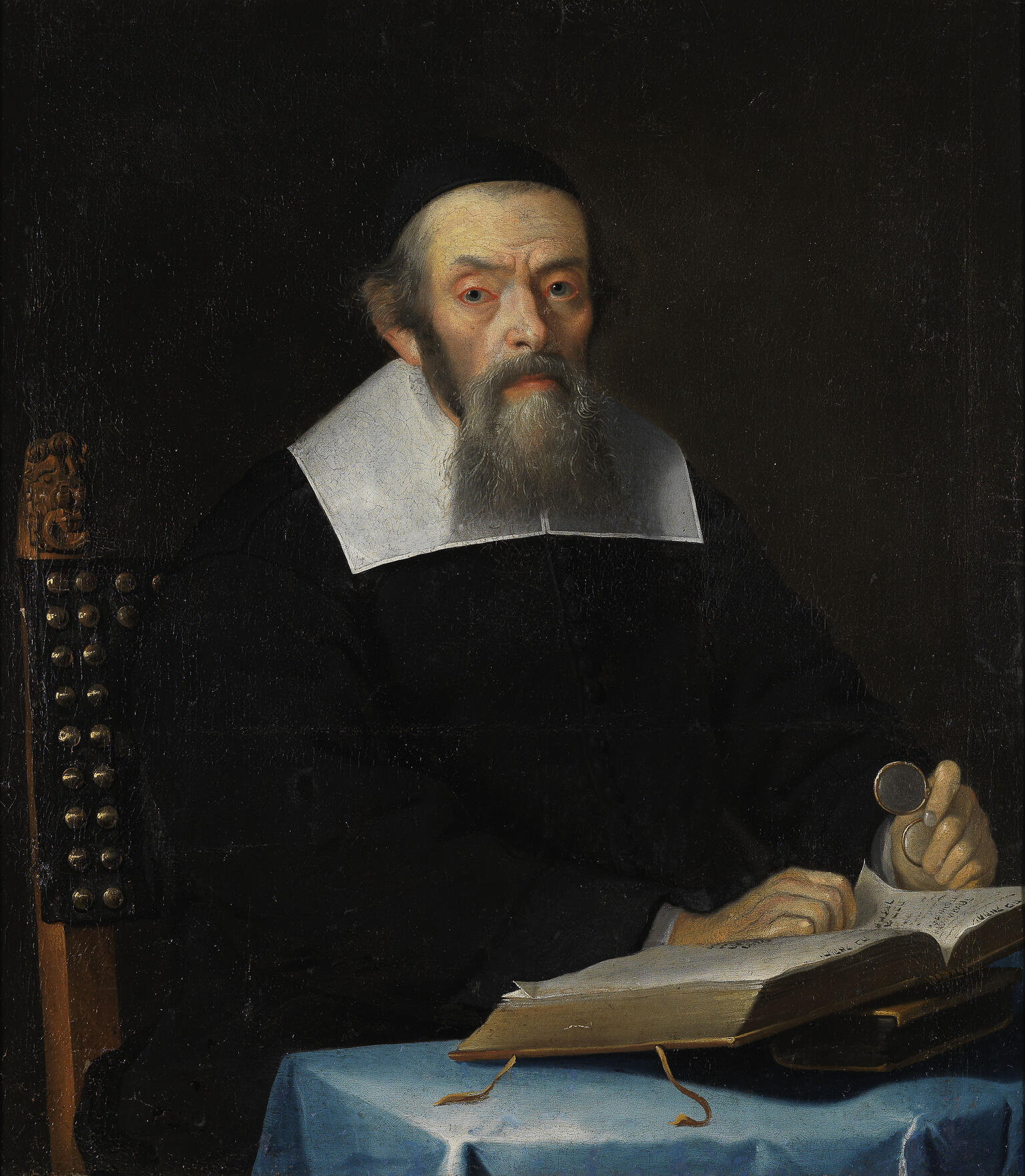 Portrait painting of man in robes with collar facing viewer and sitting at a desk, his hands resting on an open book and holding glasses in his left hand. Portrait painting of man in robes with collar facing viewer and sitting at a desk, his hands resting on an open book and holding glasses in his left hand.