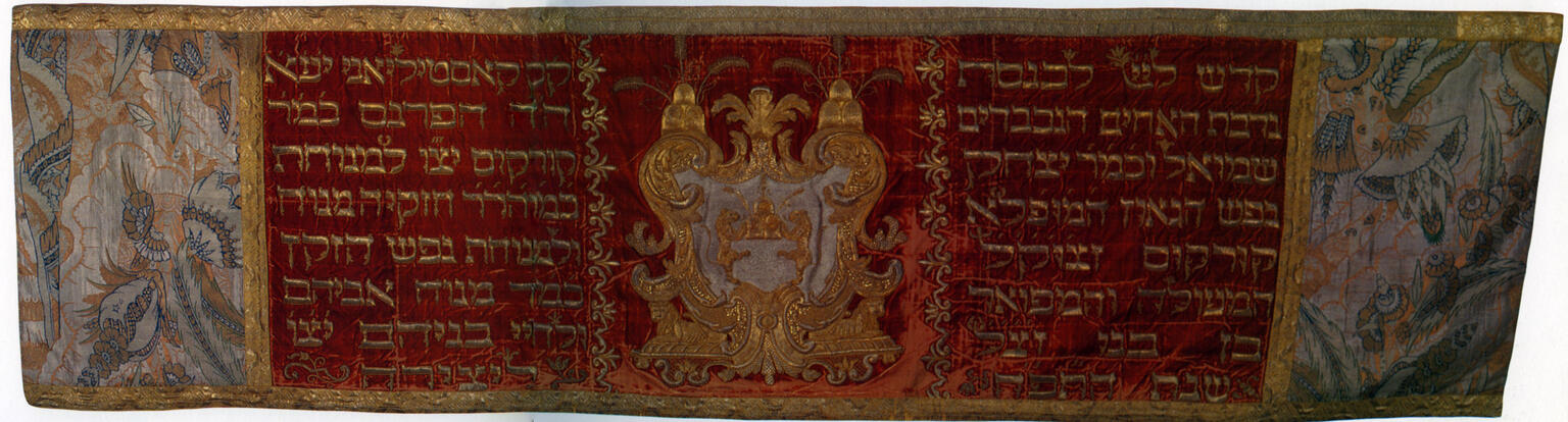 Cloth with embroidered crest in the center with Hebrew text on either side, with floral motifs on left and right edges. 