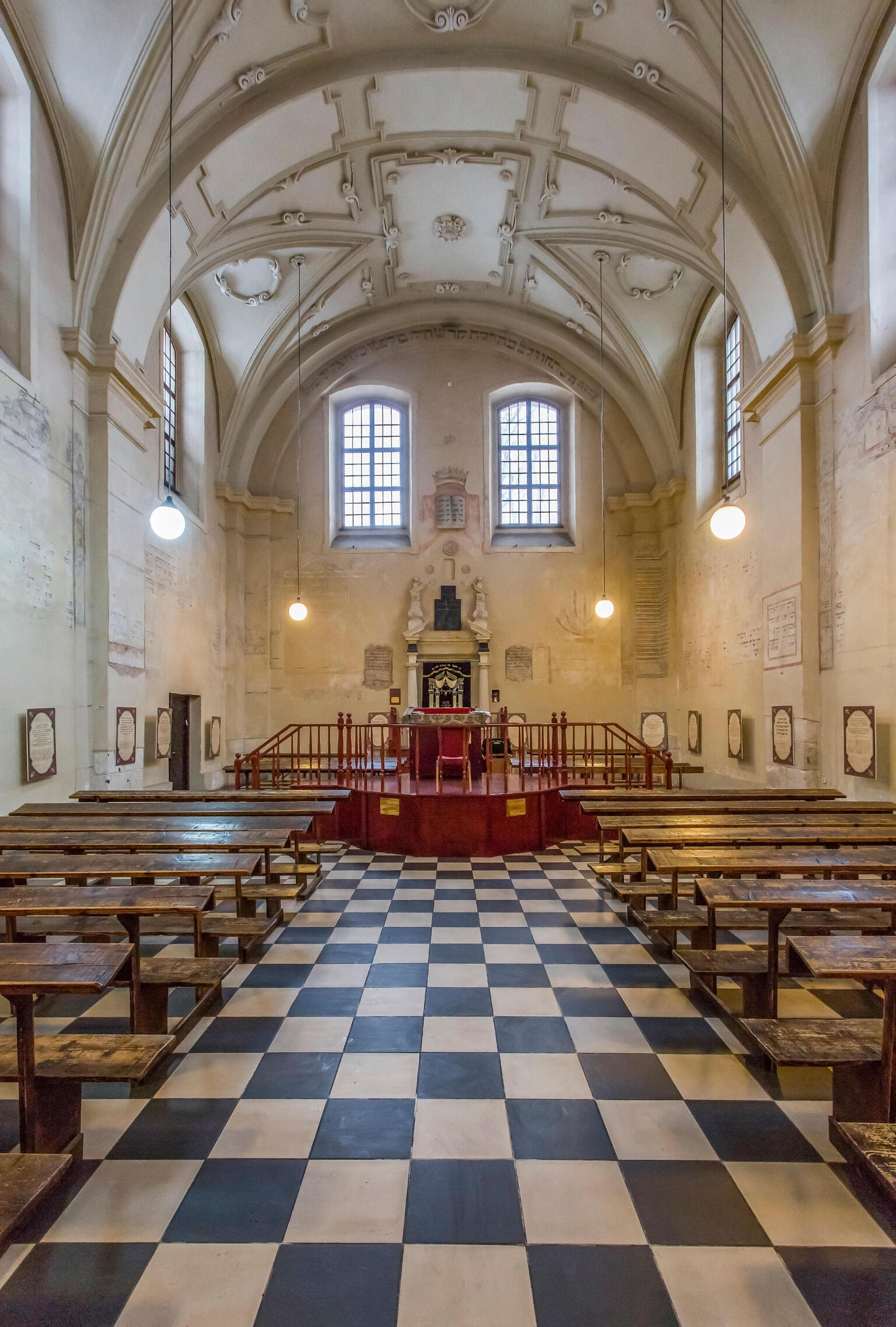 Photograph of room with vaulted ceiling, checkered floor, rows of pews, and platform with Torah ark on far wall. 