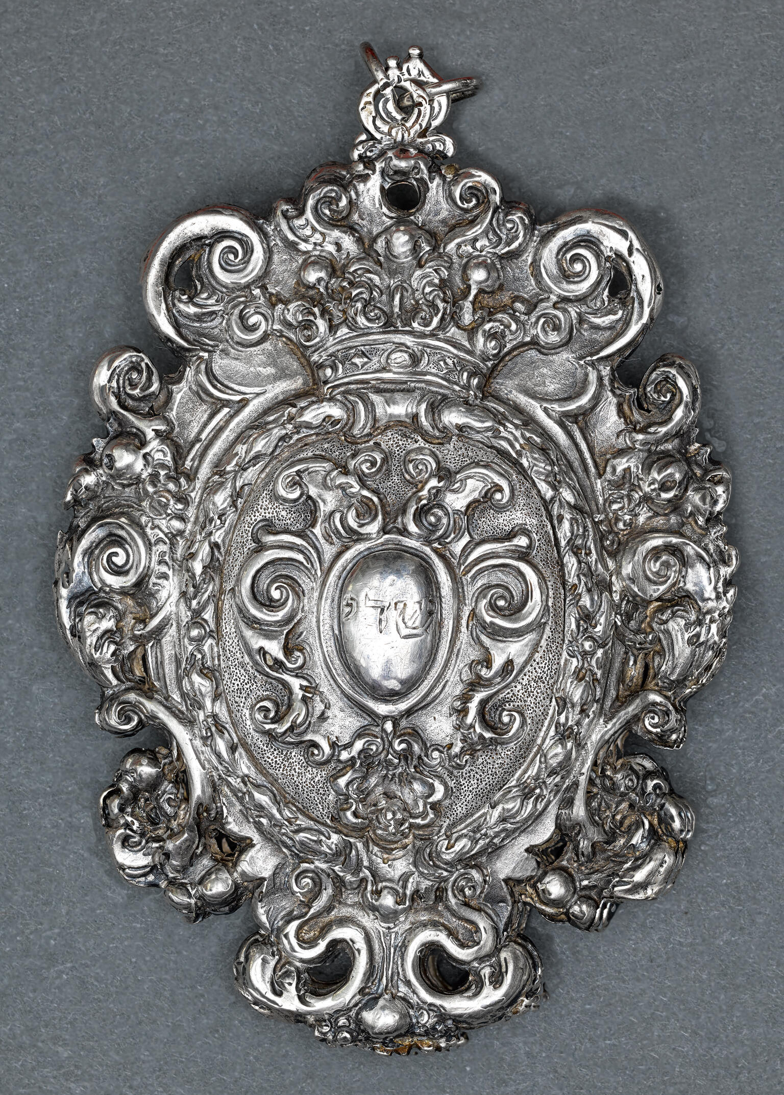 Silver amulet with decorative edges and Hebrew word etched in center. 