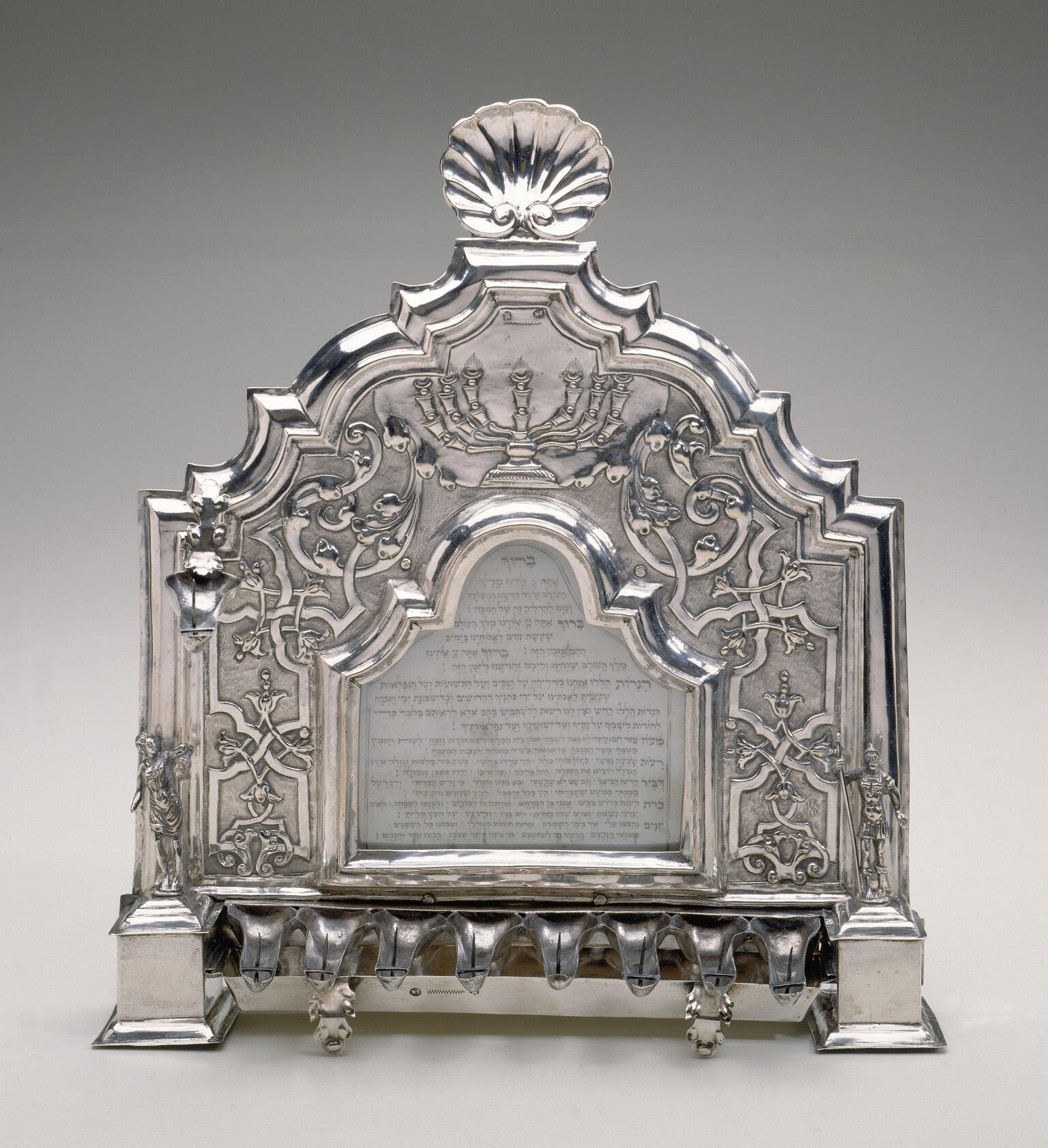 Silver oil lamp with row of eight cups below, carving of shell on top, and Hebrew inscription.
