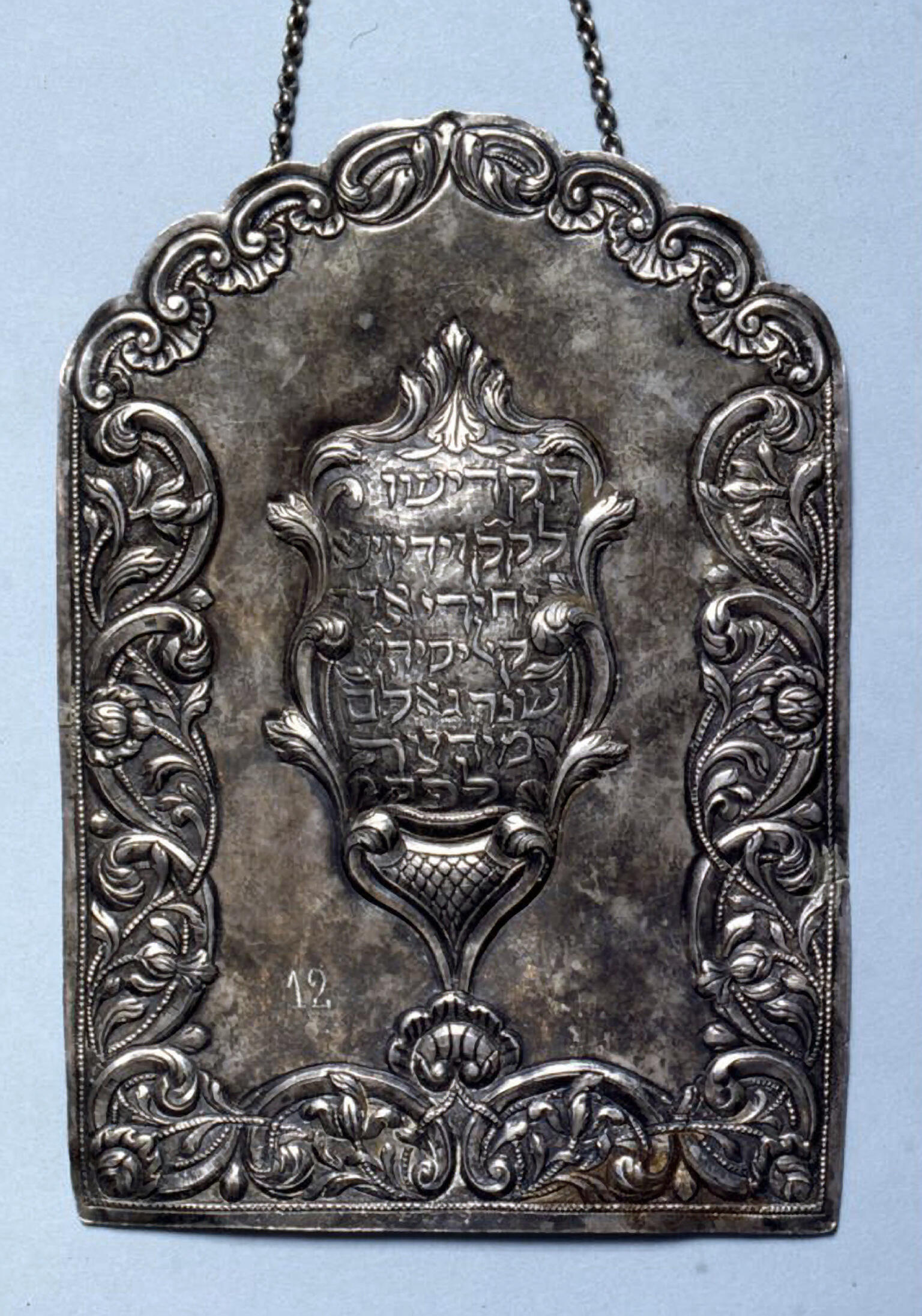 Silver shield of decorative border with Hebrew text in center.