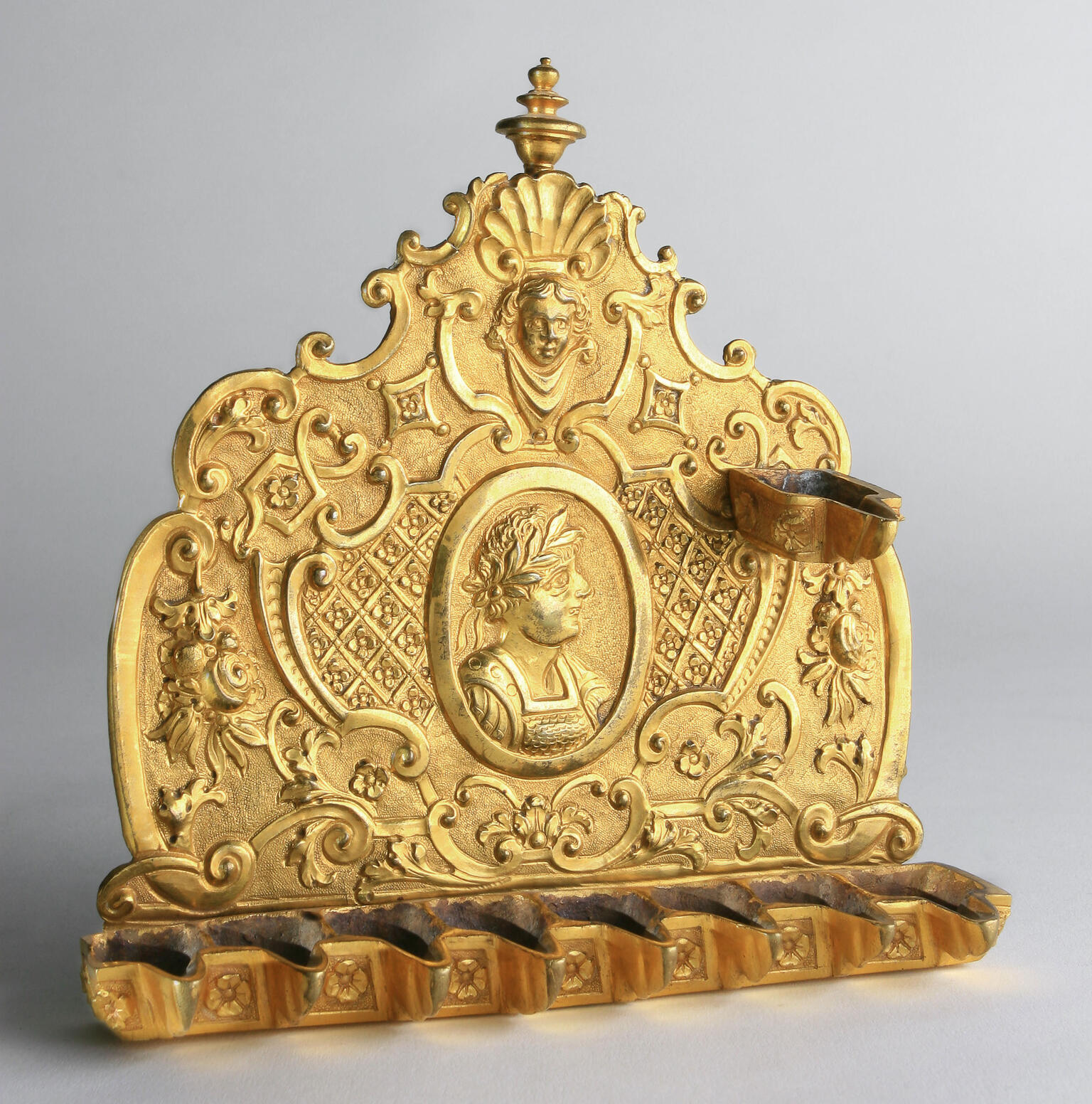 Oil lamp in the form of a shield with spouts at the bottom, of decorative motifs and profile of individual crowned with laurel wreath.