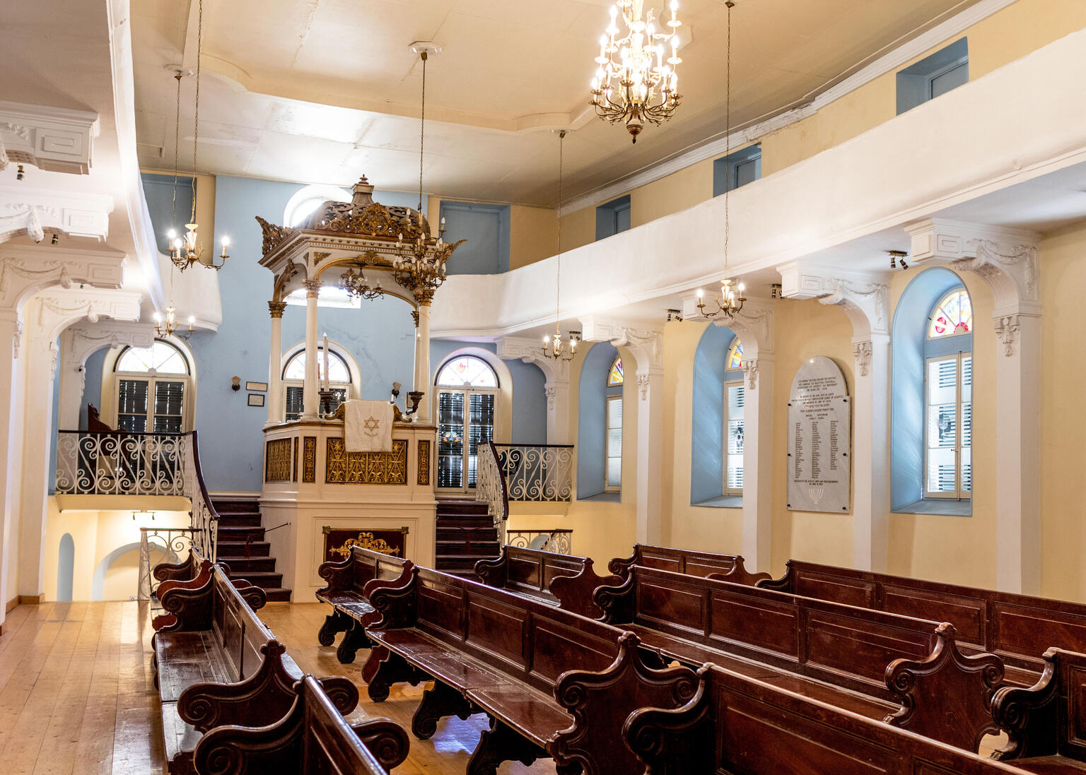 Photograph of room with high ceiling, tall podium up short staircase with golden canopy, and rows of wooden benches. 