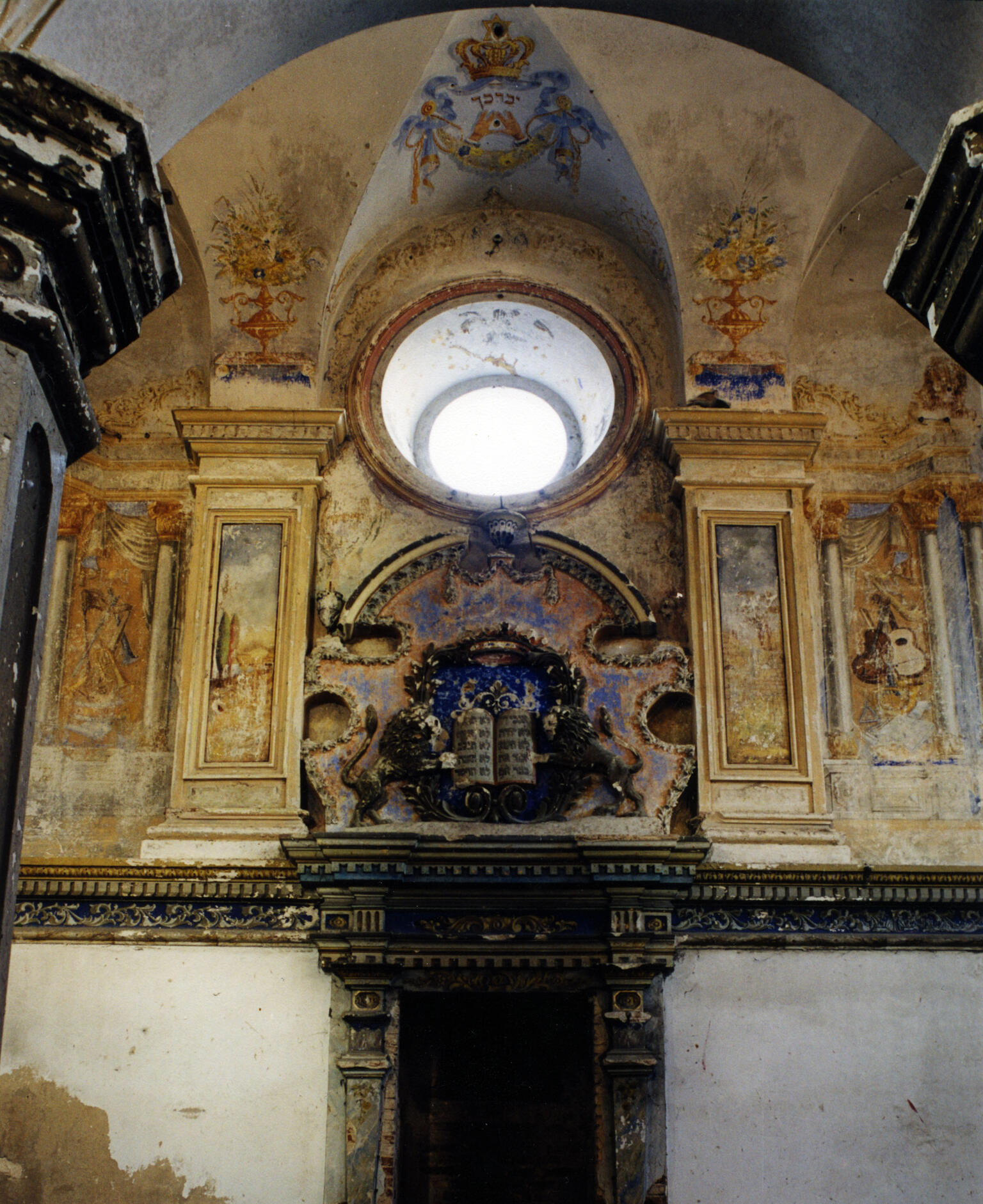 Photograph depicting round window surrounded by mural with crowns, musical instruments, and floral patterns, and sculpture of lions and tablets over doorway. 