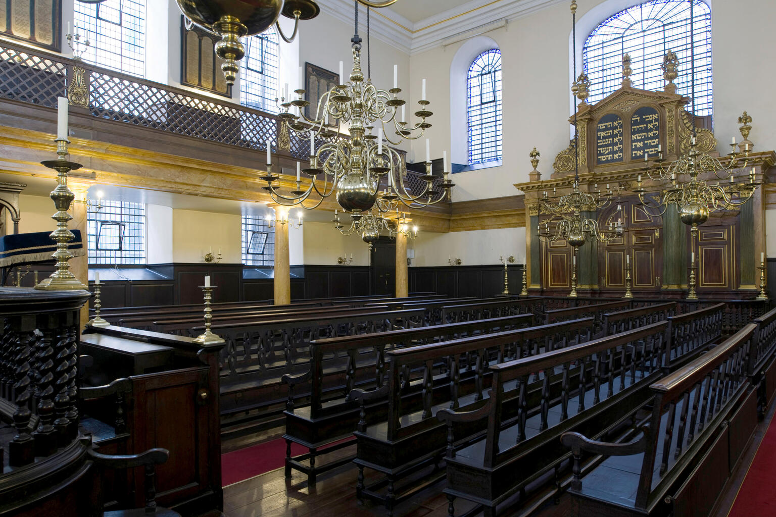 Photograph of room with wooden pews, ornate Torah ark in far wall, and second story balcony.