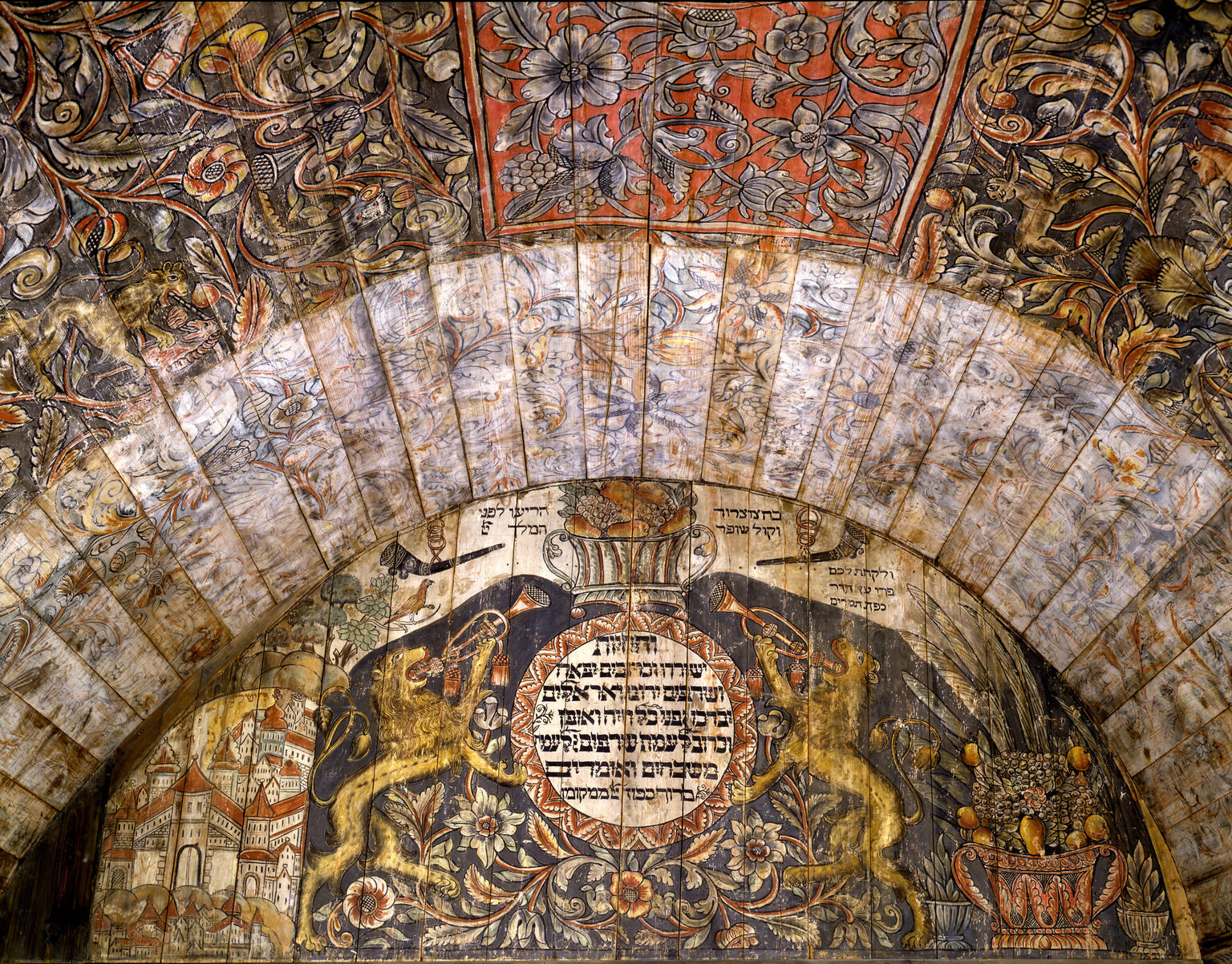Close up photograph of wall and ceiling with painting of lions on either side of Hebrew text, city on left side, cornucopia on right side, and floral and plant designs on ceiling. Close up photograph of wall and ceiling with painting of lions on either side of Hebrew text, city on left side, cornucopia on right side, and floral and plant designs on ceiling.