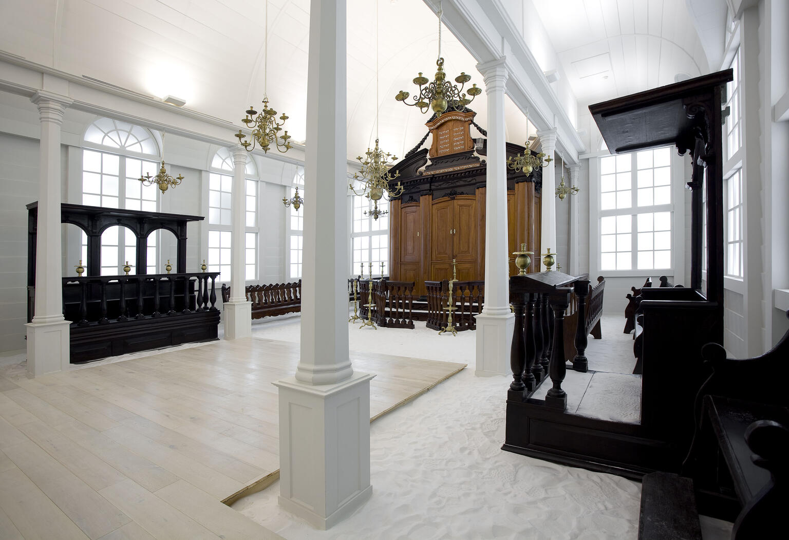 Photograph of rectangular room with arched windows, floor covered in sand, wooden cabinet on far wall, wooden benches on left side and along walls, and wooden lectern on right side. 