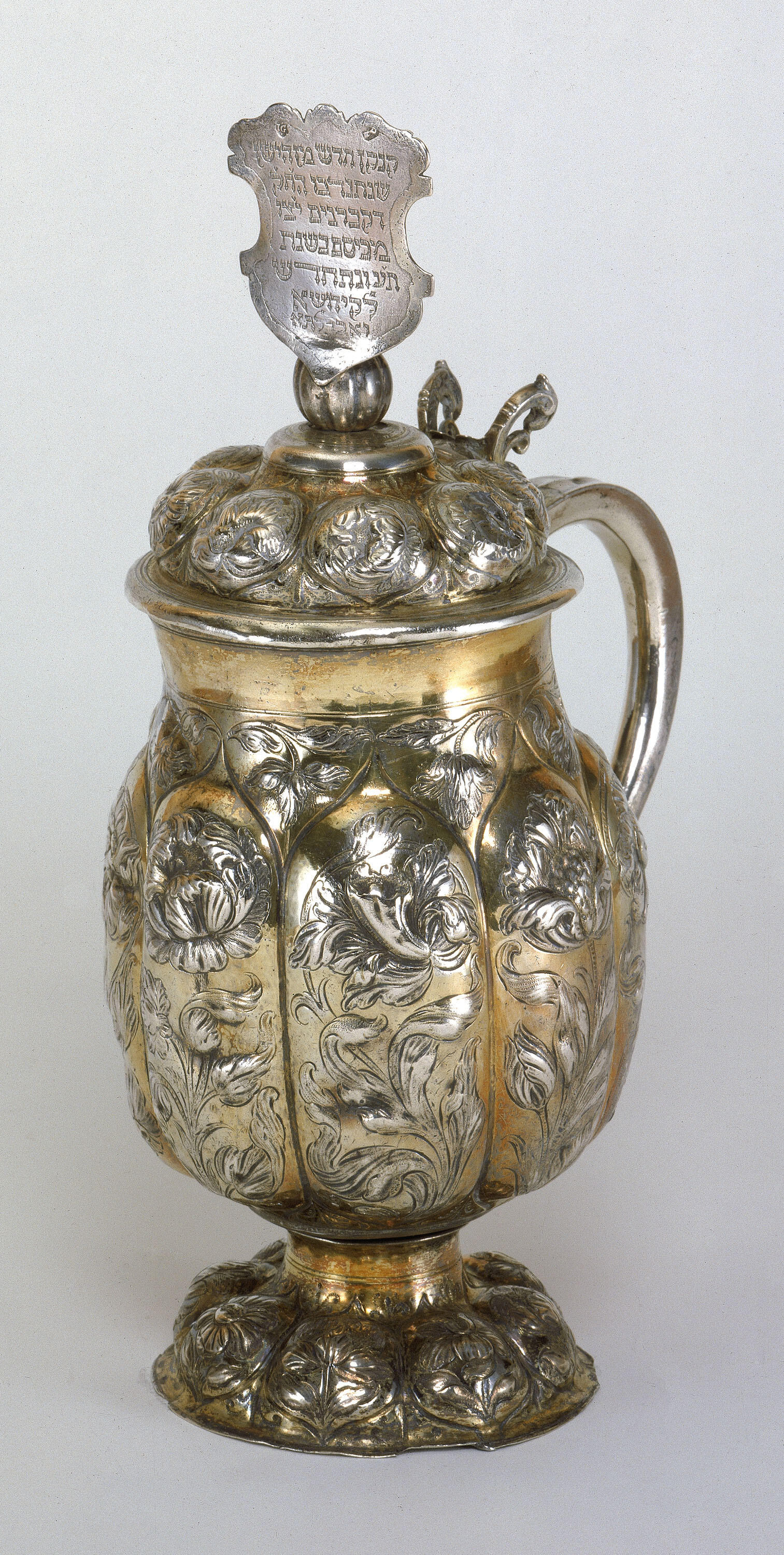 Silver pitcher with floral shapes and inscribed designs, and shield on lid with Hebrew inscription. 