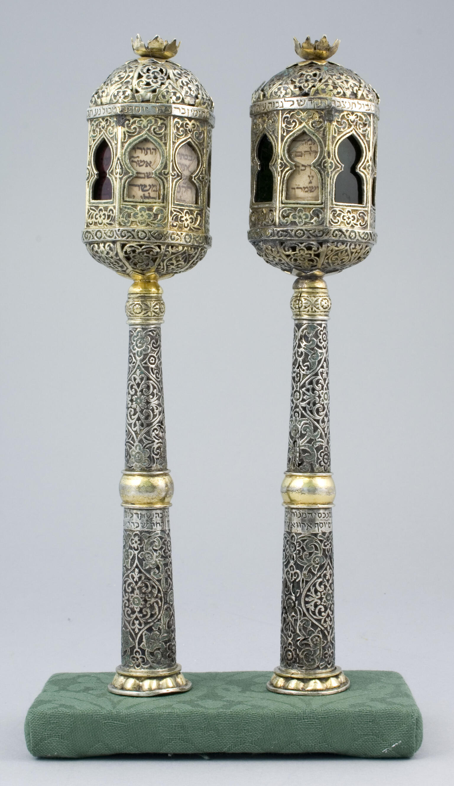 Two finials engraved with designs on the base and minaret-shaped tops with Hebrew text visible through cut-out windows. 