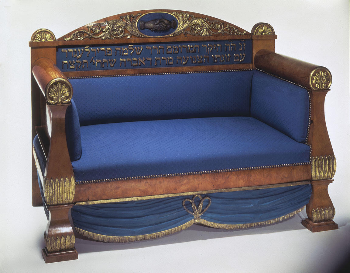 Wooden sofa with upholstered seat, featuring an image of clasped hands and Hebrew inscription on seat back. Wooden sofa with upholstered seat, featuring an image of clasped hands and Hebrew inscription on seat back.