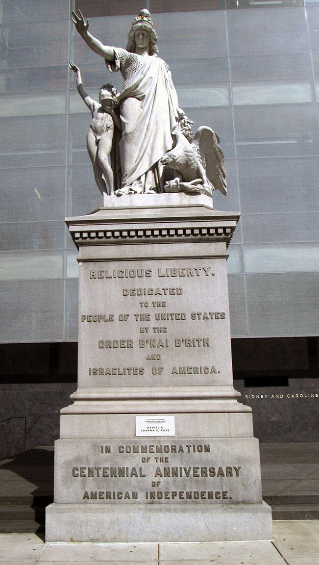 Marble monument depicting a figure dressed in coat and mantle with shield with American flag in relief on breast, with a naked boy under her right arm holding a lamp, and an eagle. 