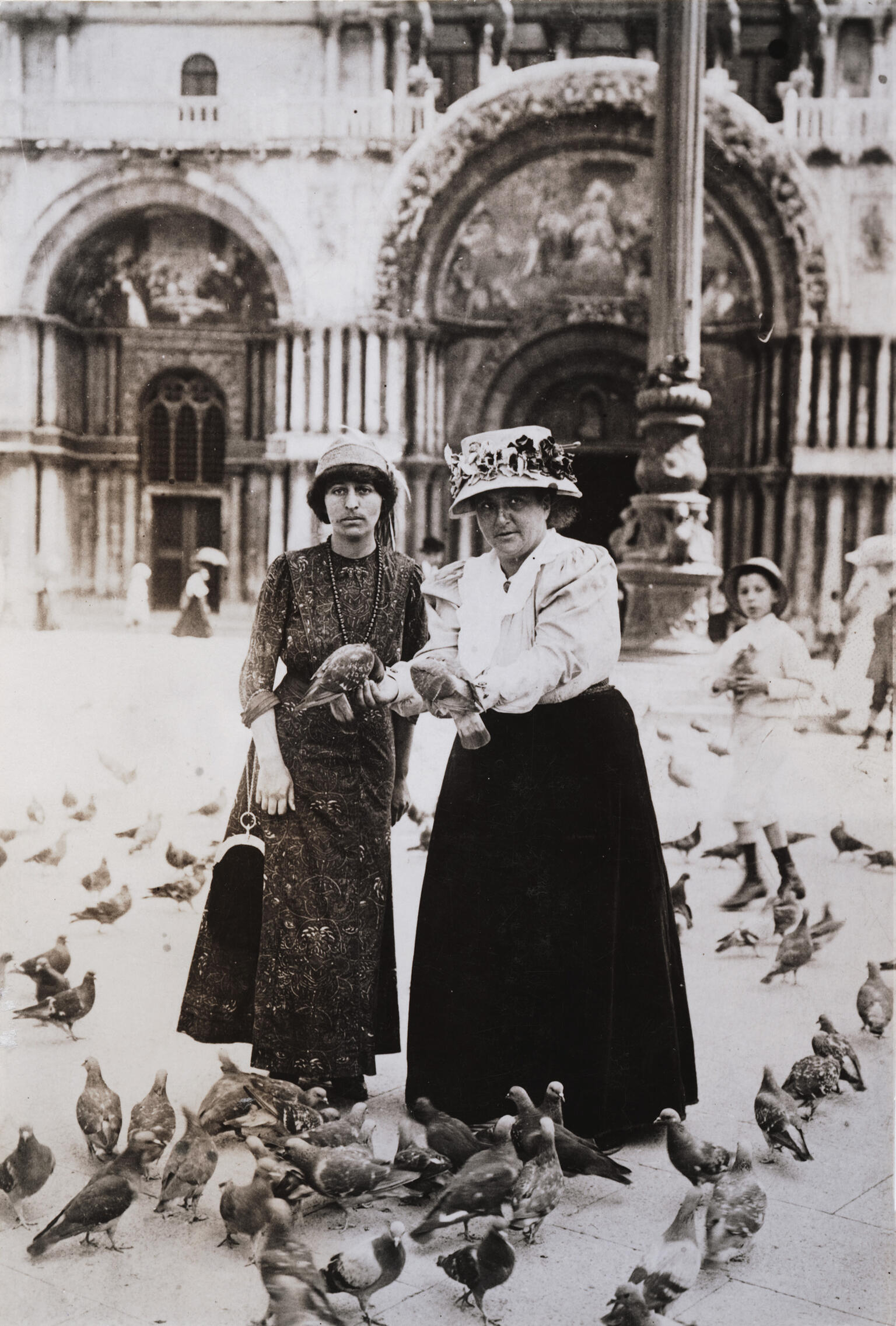 Two women in hats surrounded by pigeons in city plaza, with ornate doorway behind them.