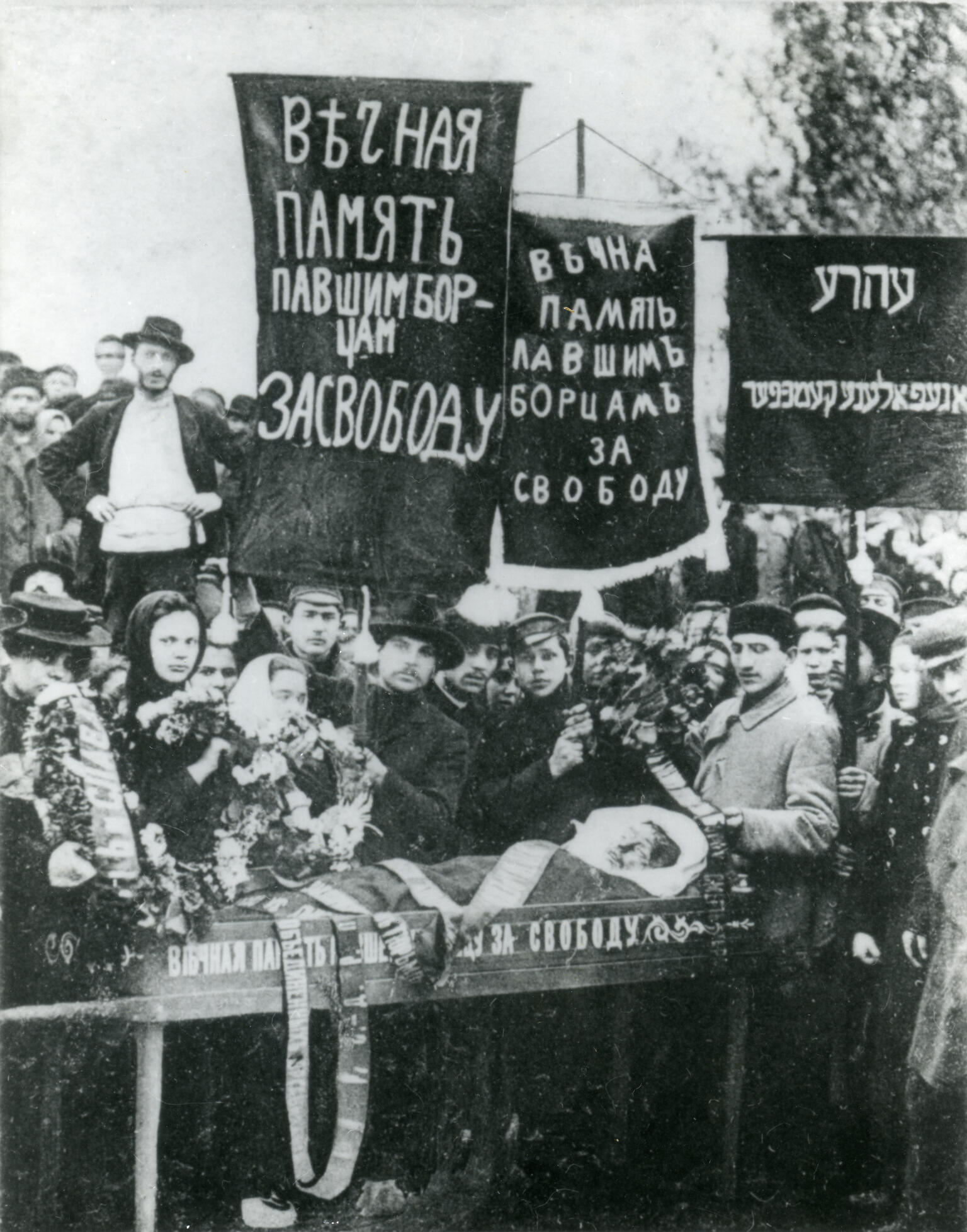 Photograph of crowd of people holding banners in Russian and Yiddish above a coffin.