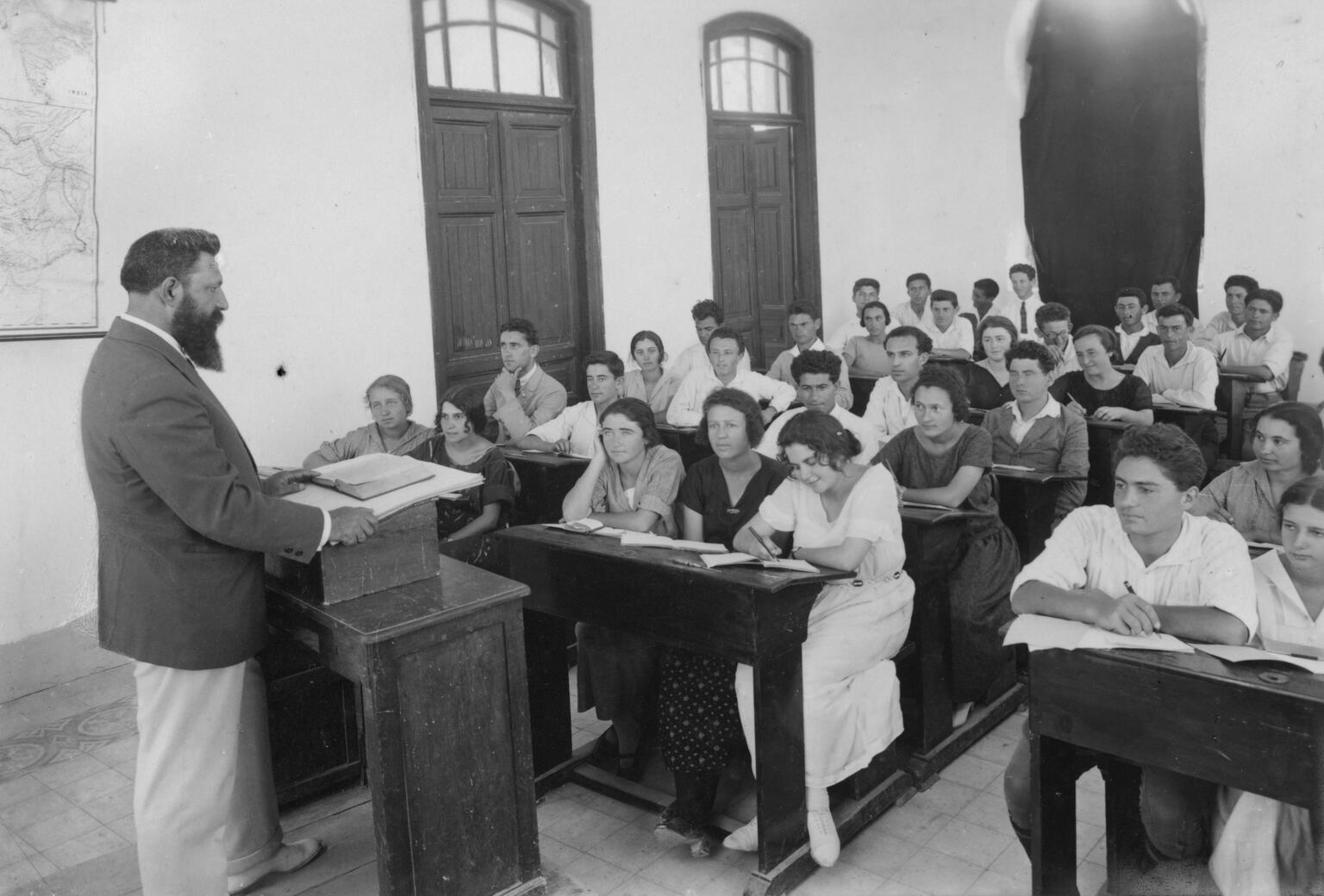 Photograph of classroom with teacher at podium and men and women at rows of desks.