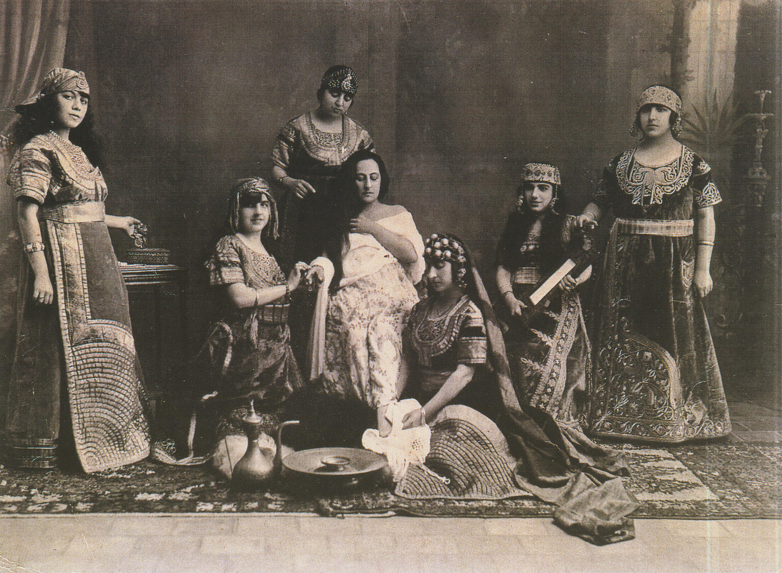 Photograph of several women in dresses and headdresses with rug and cooking implements on the floor.