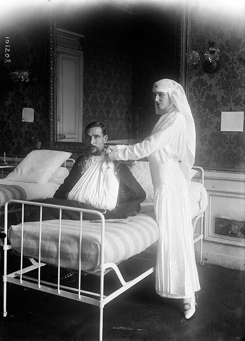 Photograph of woman in veil standing next to bed where man sits with arm in sling. Photograph of woman in veil standing next to bed where man sits with arm in sling.