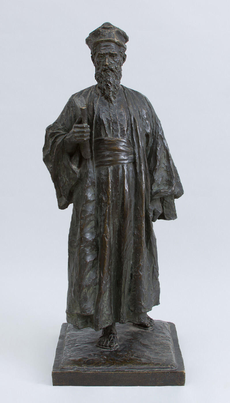 Sculpture of a figure in robe and hat, carrying a book.