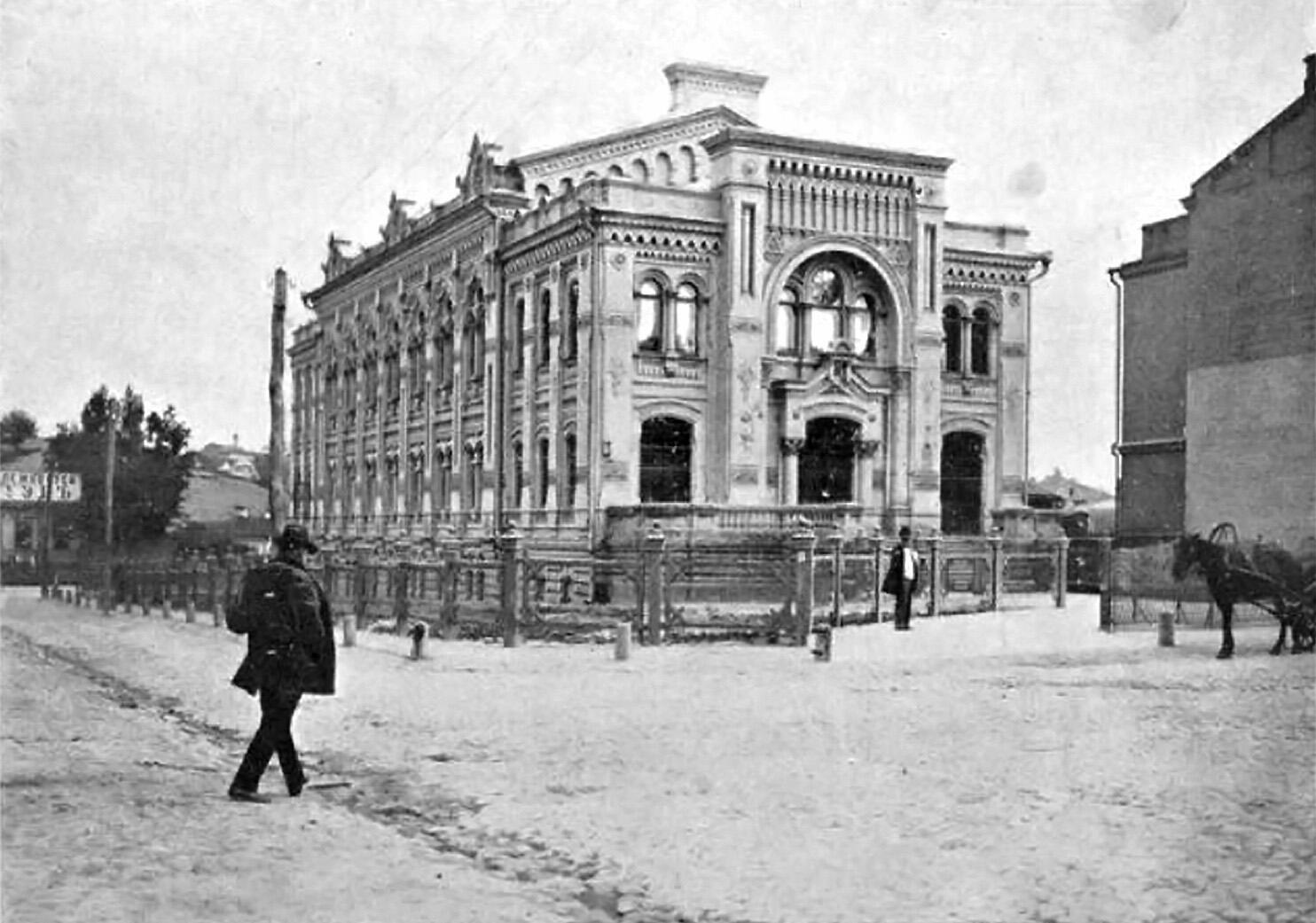 Photograph of exterior of building with rounded window frames and decorative columns, and man in hat walking toward it.