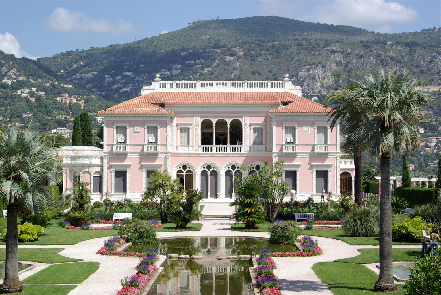 Photograph of house surrounded by gardens and fountain and flanked by two tall palm trees, with mountains in background.
