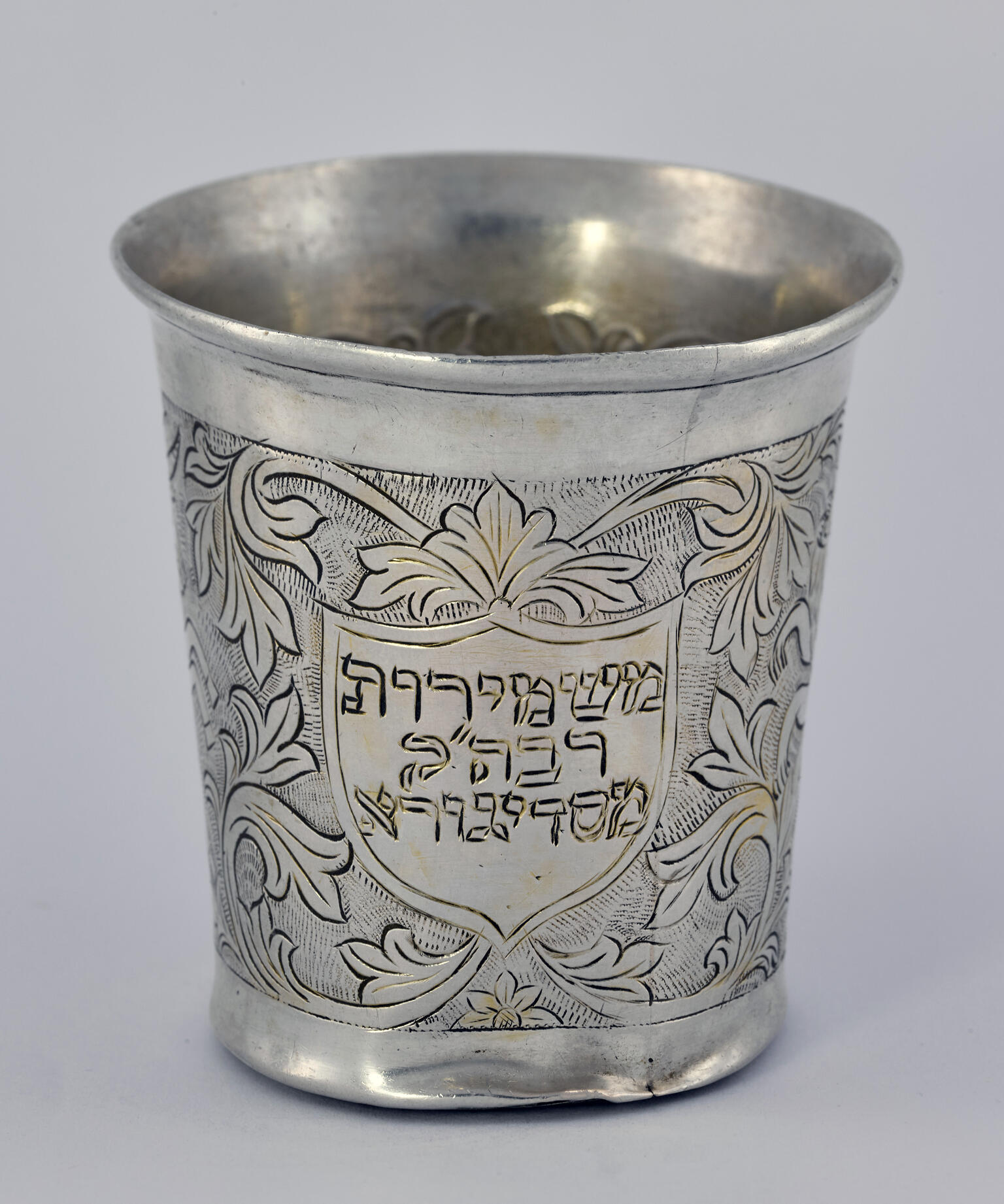 Silver cup etched with flowers, vines, and Hebrew letters in center.