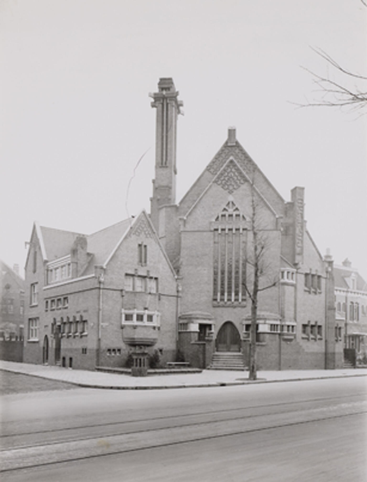 Photograph of a building with triangular roof sections, many long windows, and tower on the corner of a street.