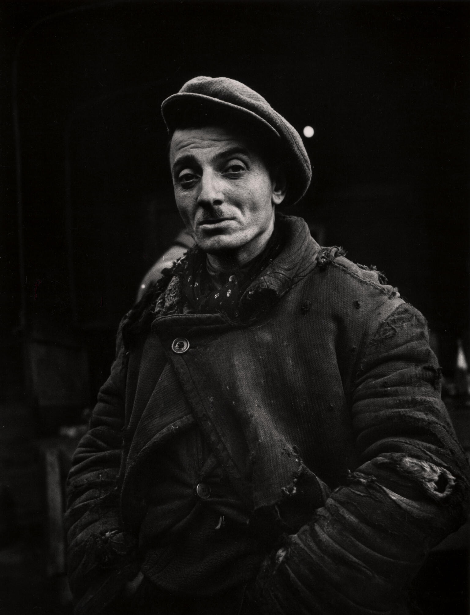 Photograph of man with mustache and hat wearing torn clothing with hands on hips, looking at camera.