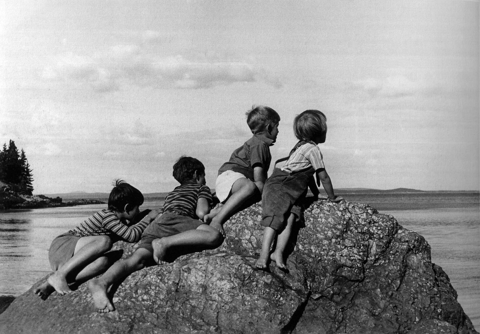 Photograph of four boys perched on a rock overlooking water and facing away from viewer. Photograph of four boys perched on a rock overlooking water and facing away from viewer.