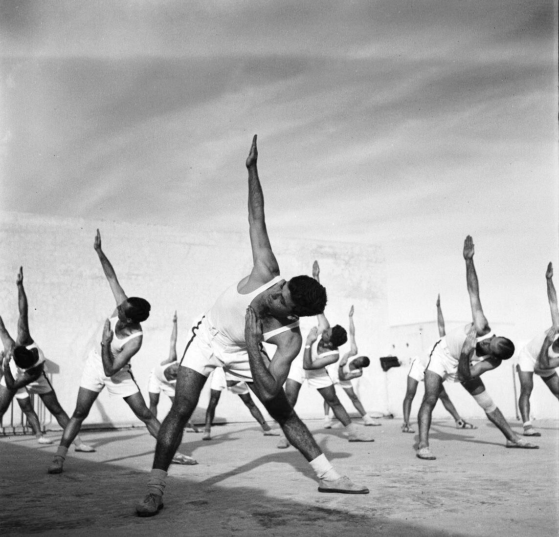 Photograph of rows of men in athletic clothing bending at the waist with their right arms pointed up and their left arms bent in an angle toward their face.