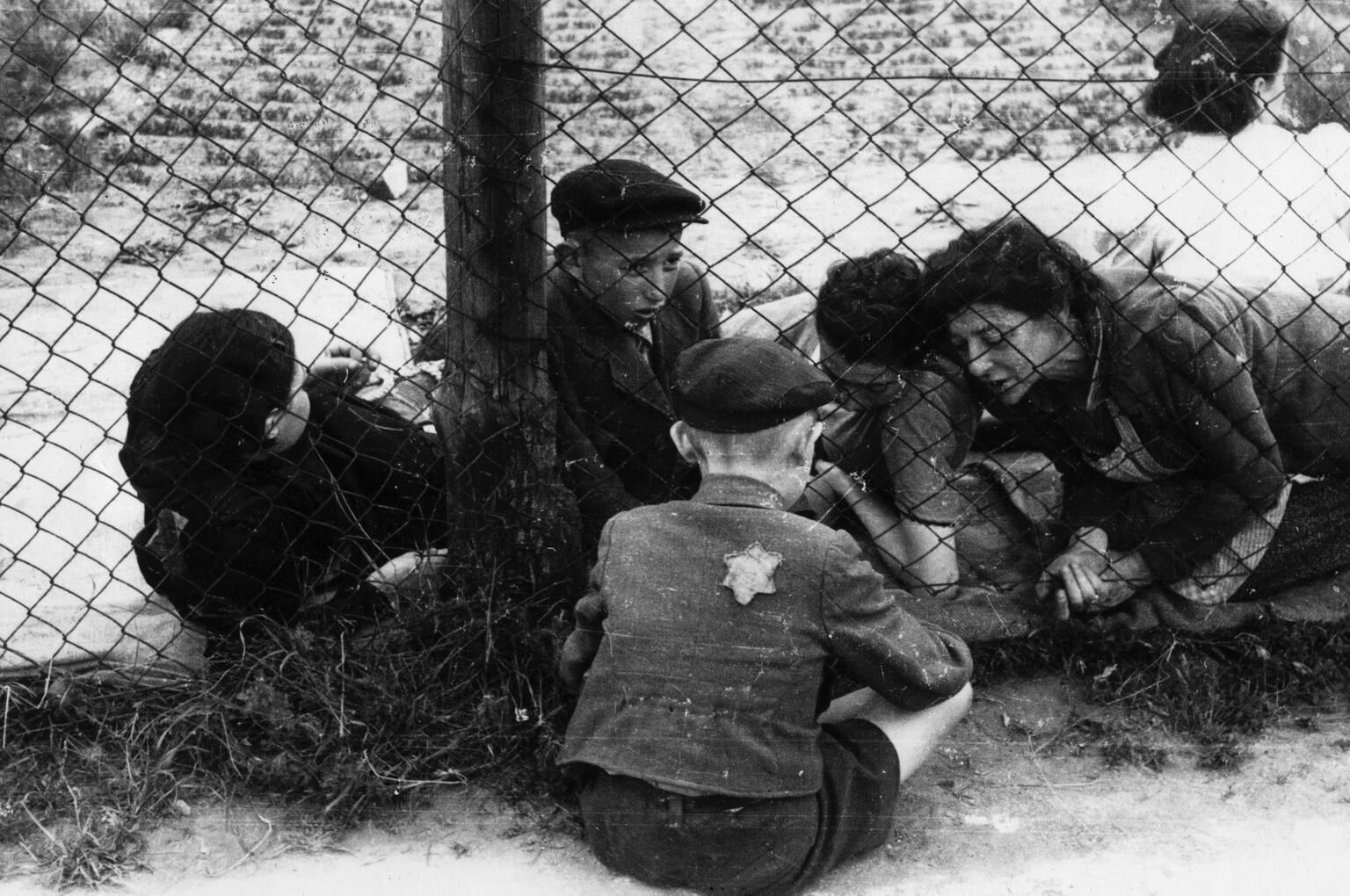 Photograph of women and children in ghetto facing photograph on the far side of a fence, interacting with another child on opposite side of fence with Star of David badge on his back.