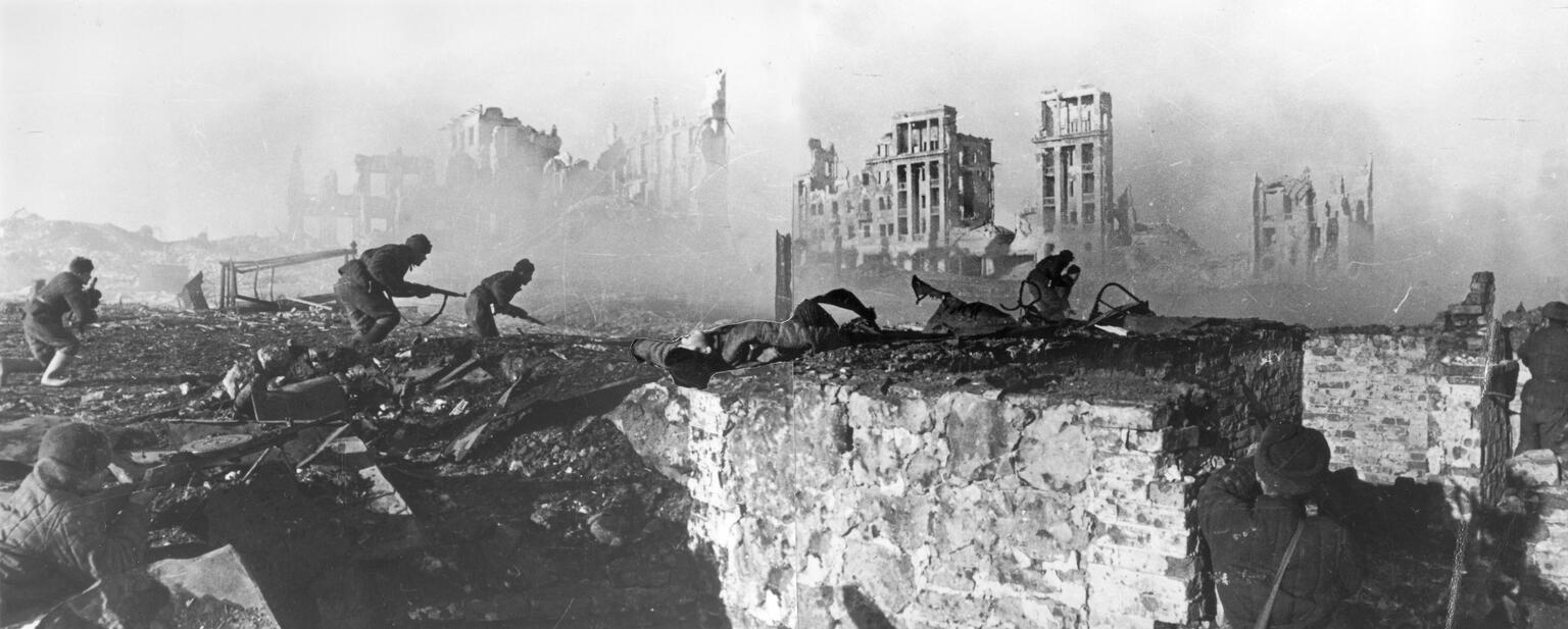 Photograph of soldiers holding guns as they approach ruins of a bombed city. 