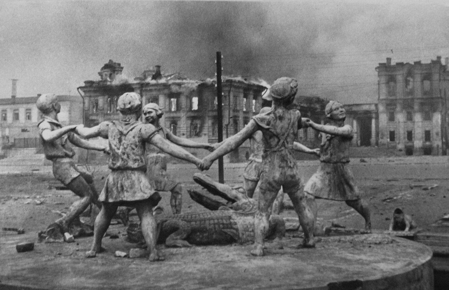 Photograph of children holding hands in a circle and dancing around an alligator statue as a city burns in the background.