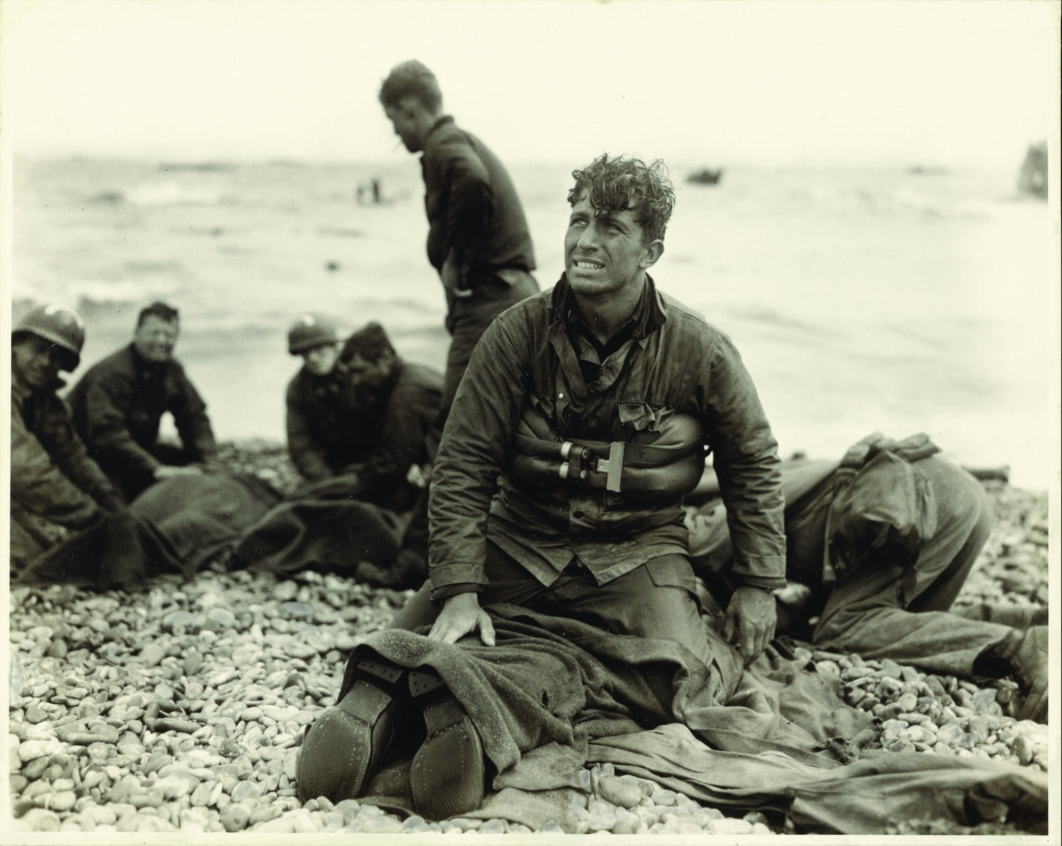 Photograph of soldiers on the beach, with man in center facing the viewer and looking up, as he kneels on top of a prone soldier.