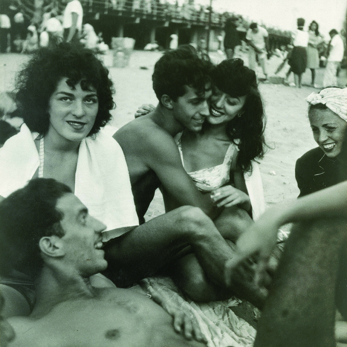 Photograph of five people sitting closely together, with one lying down, in a circle on the beach. 