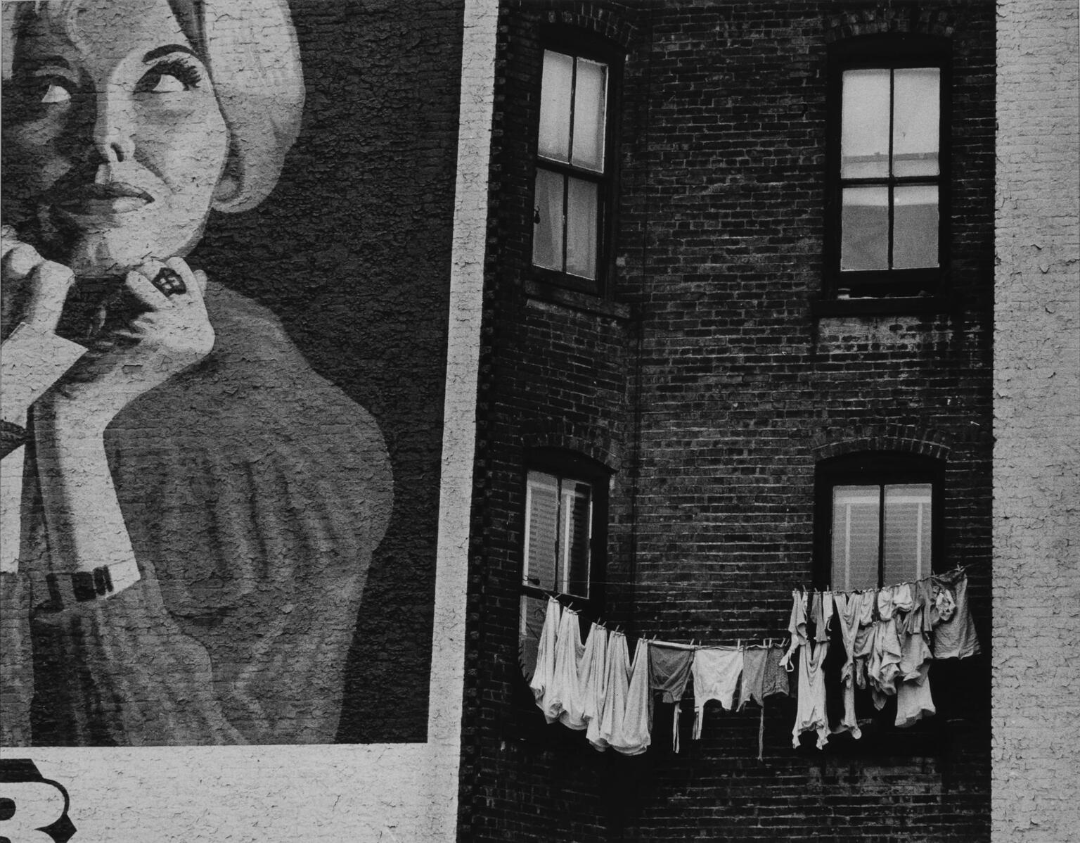 Photograph depicting painted advertisement of a woman on wall of brick building next to a clothesline on adjacent wall.