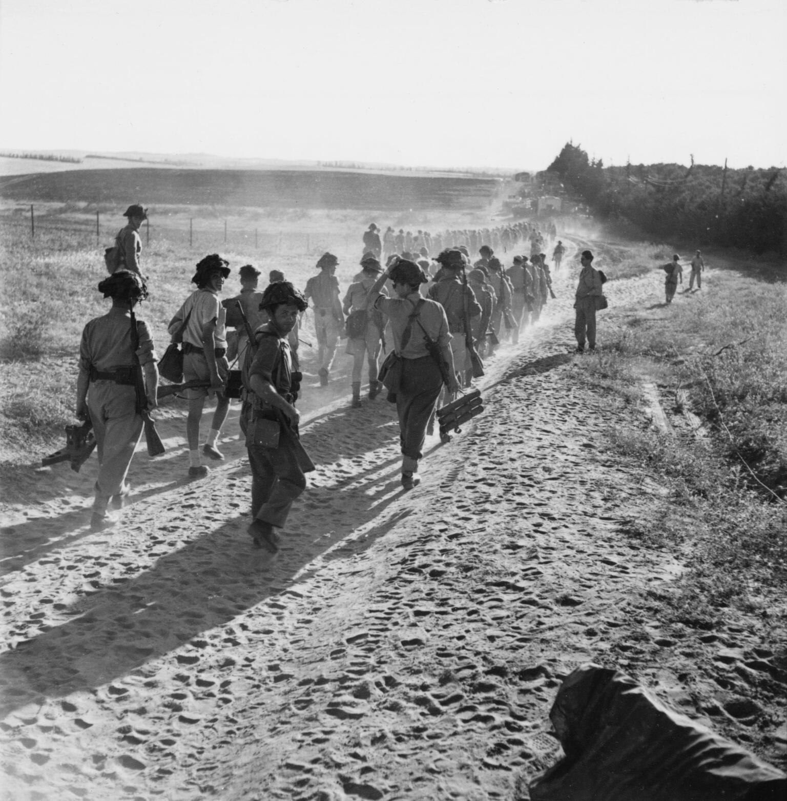 Photograph of troops marching away from the viewer on a sandy trail. Photograph of troops marching away from the viewer on a sandy trail.