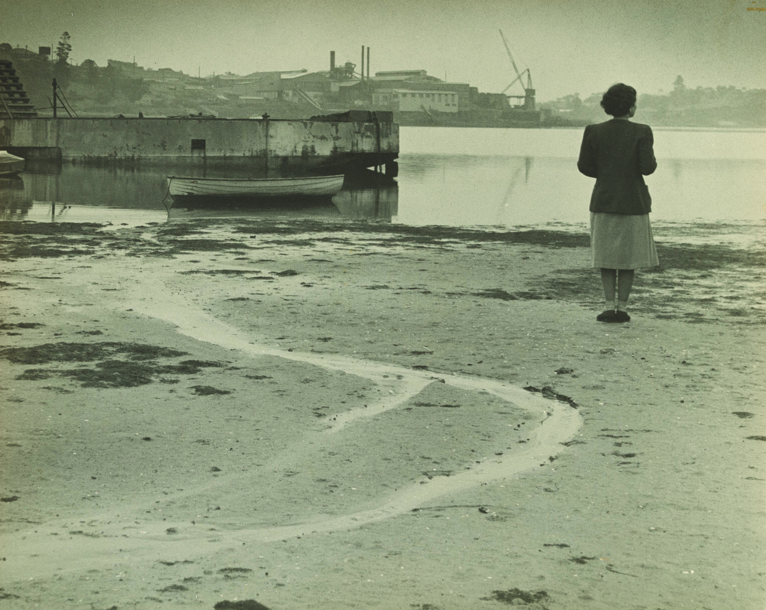 Photograph of a woman standing on a wide beach and facing away from the viewer toward a river.