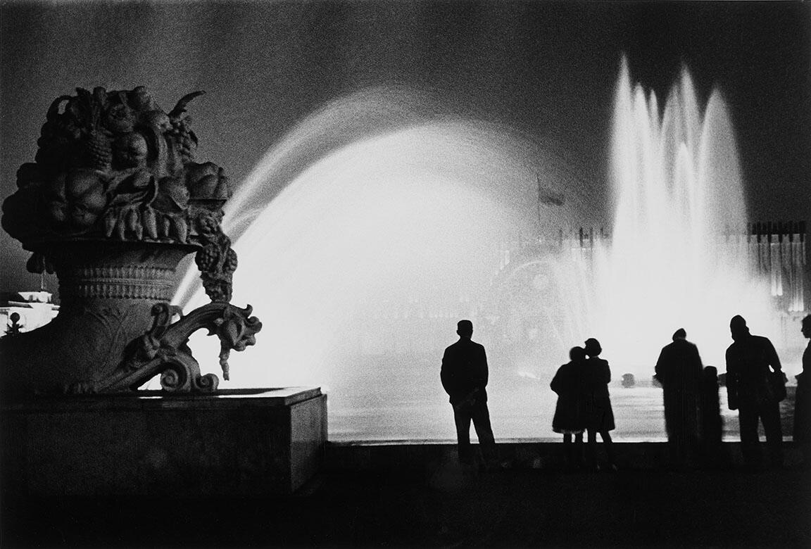 Photograph of a cornucopia statue in the left foreground silhouetted against a large fountain in the background. There are several people standing at the edge of the fountain looking away from the camera toward the water. 