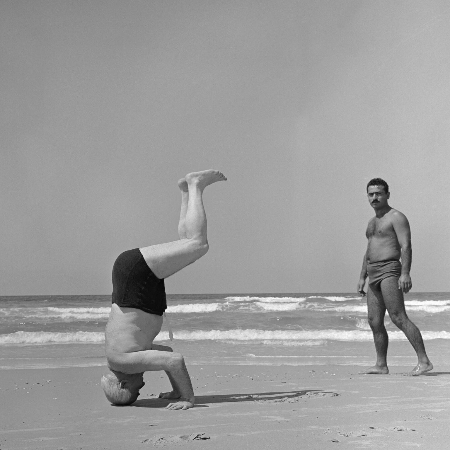 Photograph of two shirtless men on the beach, one doing a headstand with legs bent facing to the right of the viewer and the other man facing the viewer on the right side of the photograph.