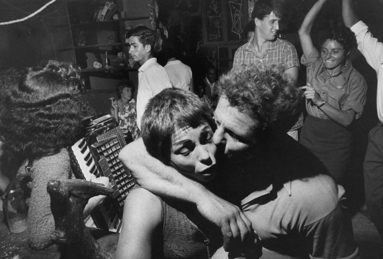 Photograph of several people socializing, including someone with accordian, three figures dancing, and a man with his arm around a woman and kissing her cheek in the foreground.