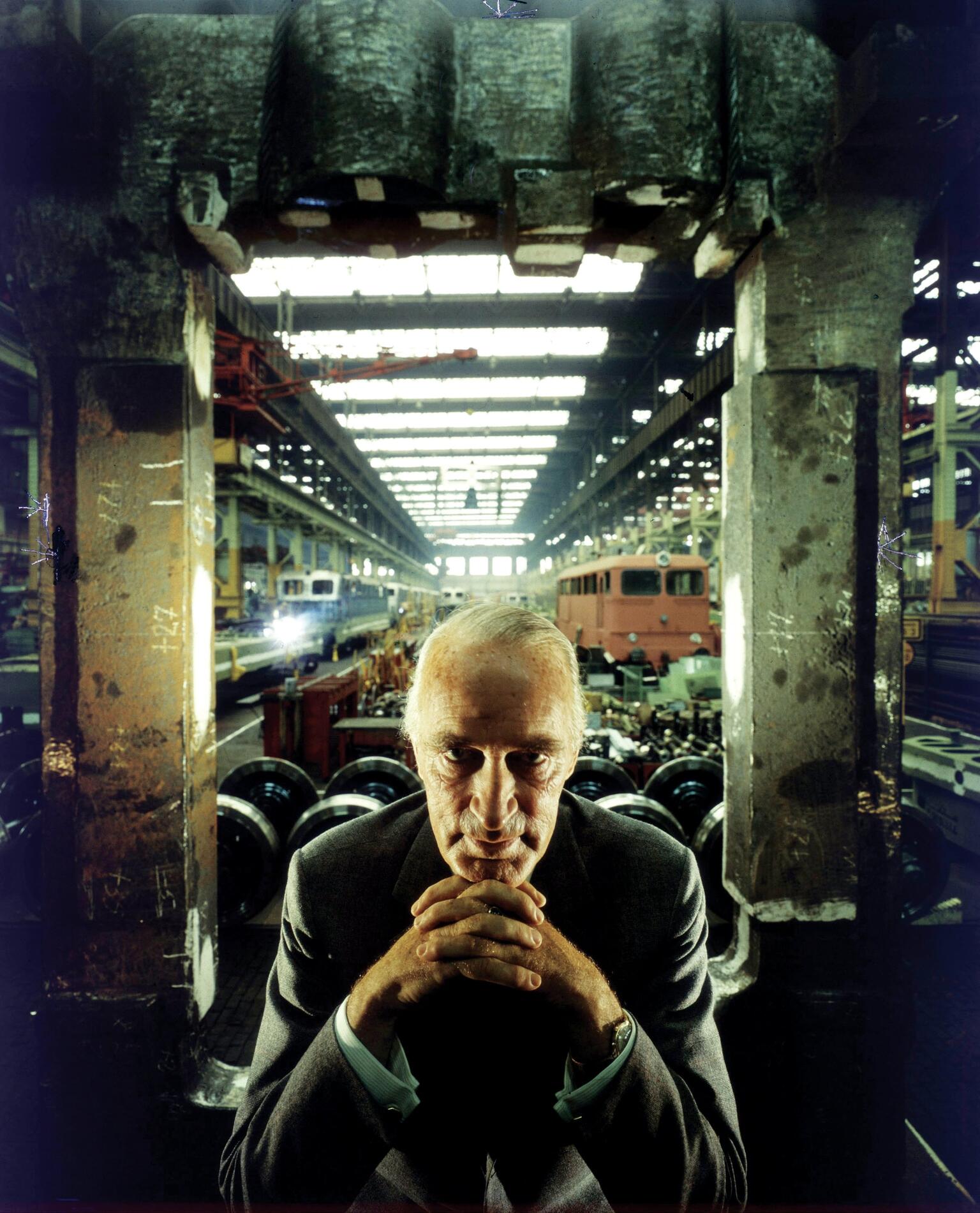 Photograph of man wearing a suit seated in warehouse facing camera with chin resting on folded hands, framed by two concrete pillars. 