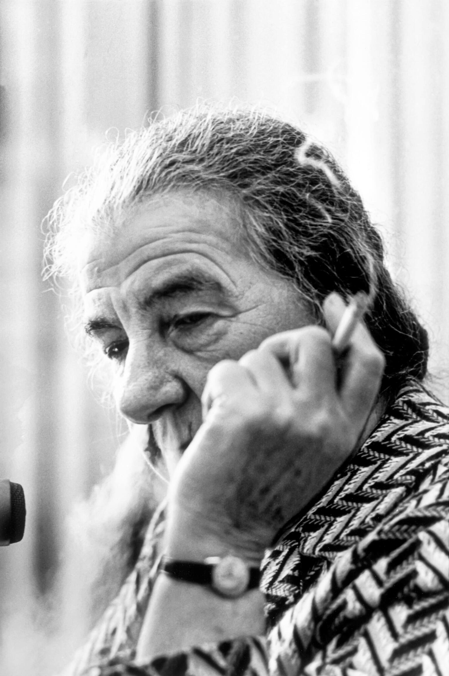 Photograph of an elderly woman smoking a cigarette, whose face is partially blocked by her hand holding the cigarette. 