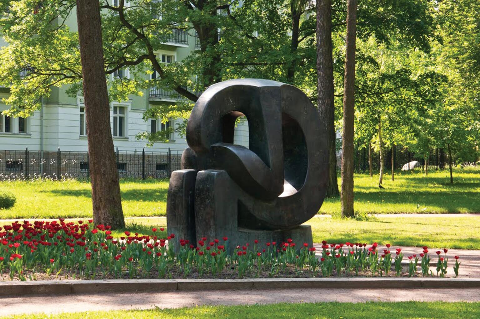 Monument depicting curved, stylized figure with head in hands set among flowers in a grassy park with a house in the background.