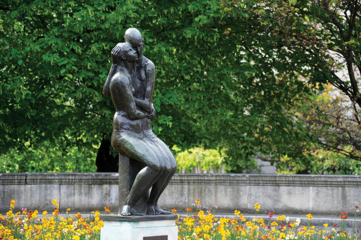 Bronze sculpture of young couple, seated side-by-side and cheek-to-cheek in a garden. The ground is covered by leaves and there are many trees in the background beyond a low wall. Bronze sculpture of young couple, seated side-by-side and cheek-to-cheek in a garden. The ground is covered by leaves and there are many trees in the background beyond a low wall.