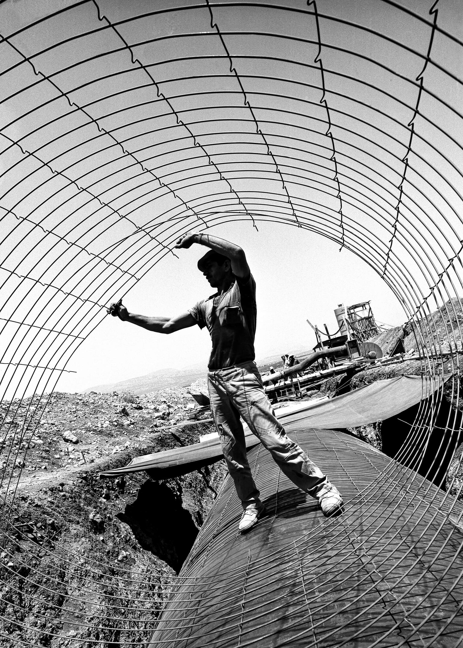 Photograph of man holding a wire cylinder and standing on a large pipe, with a construction site behind him.