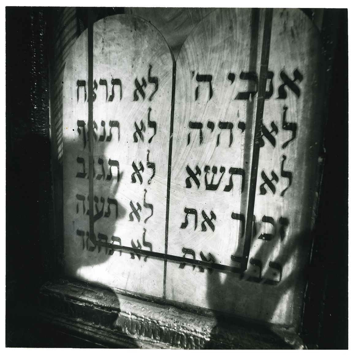 Photograph featuring shadow of a face and a hand over a wall with Hebrew text. 